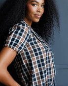A woman with long, curly black hair wearing a short-sleeve plaid button-up shirt in brown, black, and white, posing against a dark gray wall.