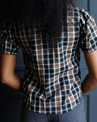 A person shown from the back wearing a short-sleeve plaid button-up shirt with a brown, black, and white check pattern, paired with gray jeans against a dark blue paneled wall.