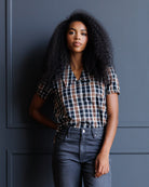 A woman with voluminous dark curly hair stands against a dark blue paneled wall, wearing a short-sleeve button-up plaid shirt in navy, white, and brown, paired with dark gray jeans.