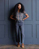 A woman with voluminous dark curly hair stands against a dark blue paneled wall, wearing a short-sleeve black, white, and orange plaid button-down shirt tucked into high-waisted, wide-leg gray denim jeans, paired with tan lace-up ankle boots.