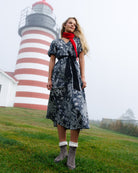 A woman stands on a grassy hill in front of a red-and-white striped lighthouse, wearing a black and white floral midi dress with a black waist tie and a bright red neck scarf, paired with gray boots.