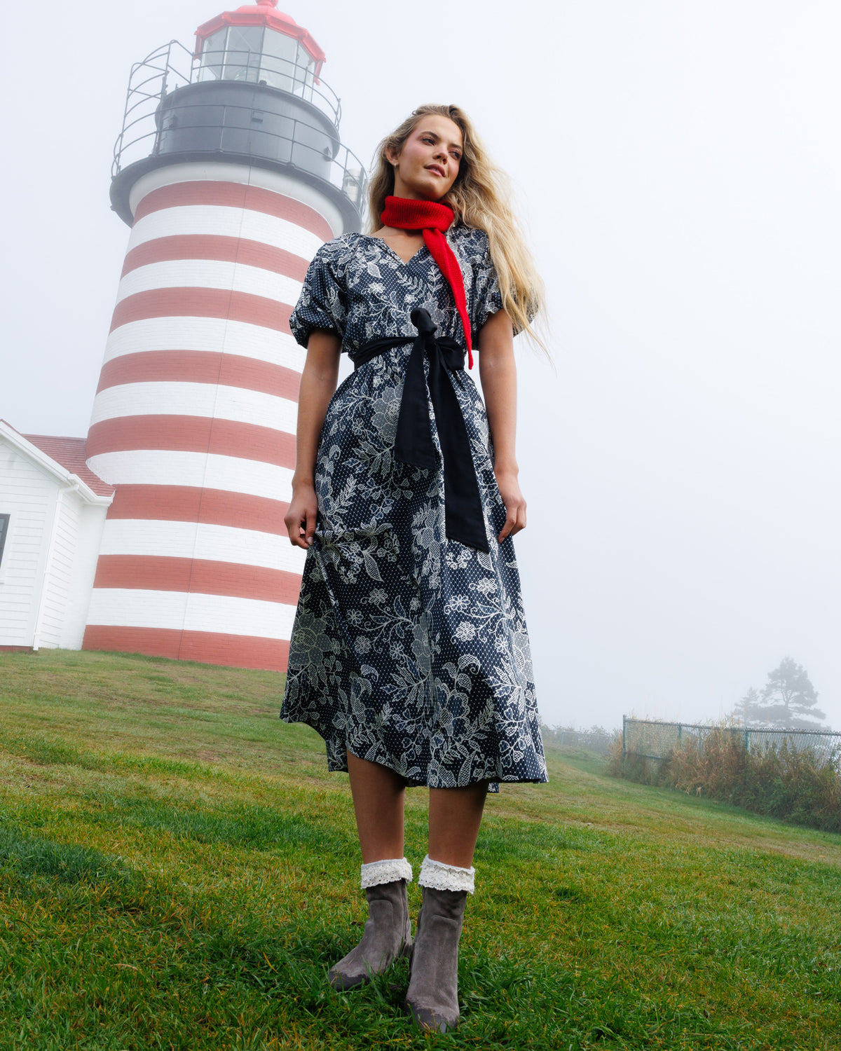 A woman stands on a grassy hill in front of a red-and-white striped lighthouse, wearing a black and white floral midi dress with a black waist tie and a bright red neck scarf, paired with gray boots.