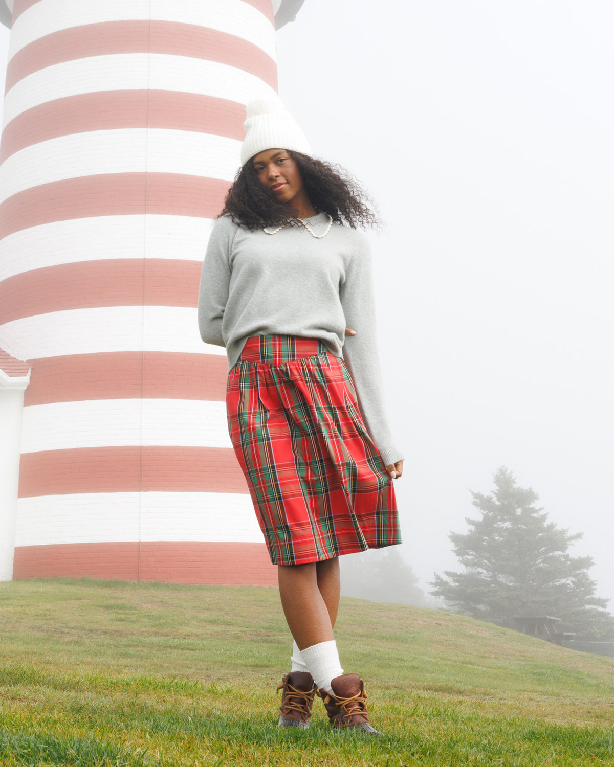 A woman stands outdoors in front of a large red-and-white striped lighthouse on a foggy day. She is wearing a light gray knit sweater, a red tartan plaid skirt, a white beanie, white socks, and brown lace-up boots.