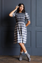 A woman models a short-sleeved plaid dress with a smocked bodice, standing against a dark blue paneled wall.