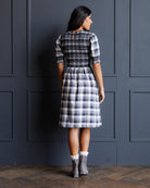 A woman stands with her back to the camera wearing a knee-length plaid dress with puff sleeves and a back keyhole detail, paired with gray socks and boots, in front of a dark paneled wall.