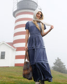 A woman wearing a navy blue tiered maxi dress and a cream-colored bonnet stands in front of a red and white striped lighthouse, holding a plaid tote bag.