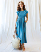 Woman wearing a blue dress holding a woven bag against a white curtain background