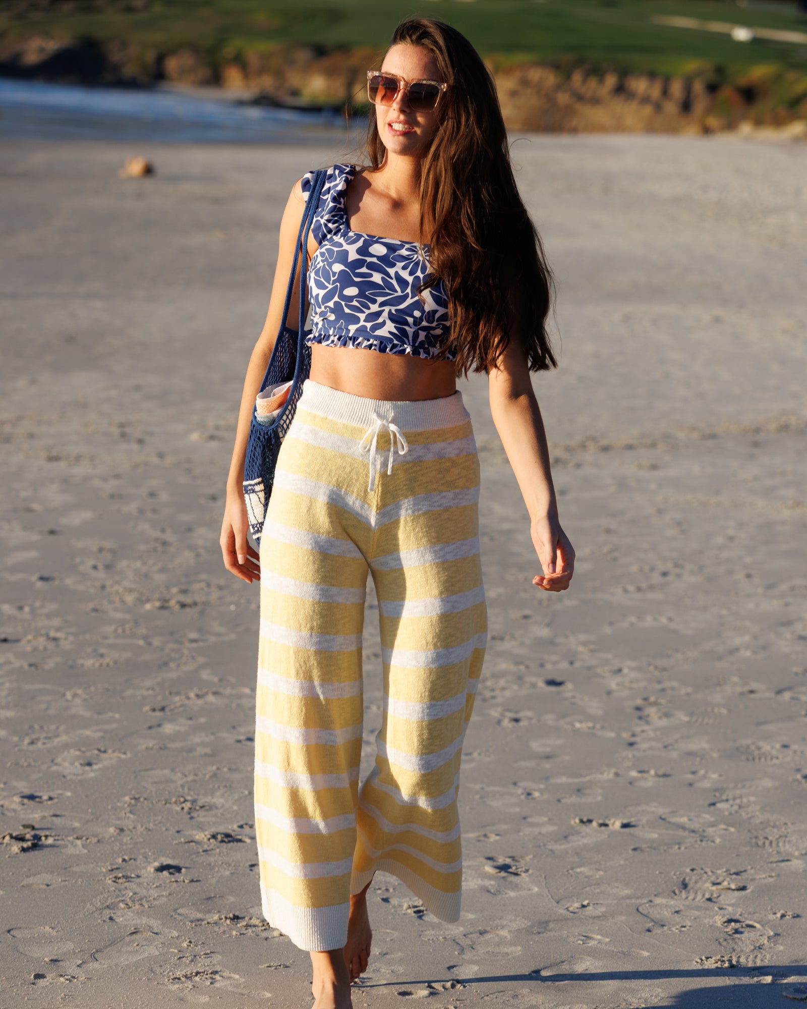 Woman in a blue and white patterned top and yellow and white striped pants walking on a beach.