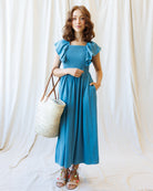 Woman in a blue dress holding a straw bag against a white curtain background
