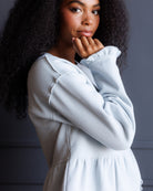 A woman with long, dark, curly hair wearing a light blue long-sleeve top, posing with her hand on her chin against a dark gray background.