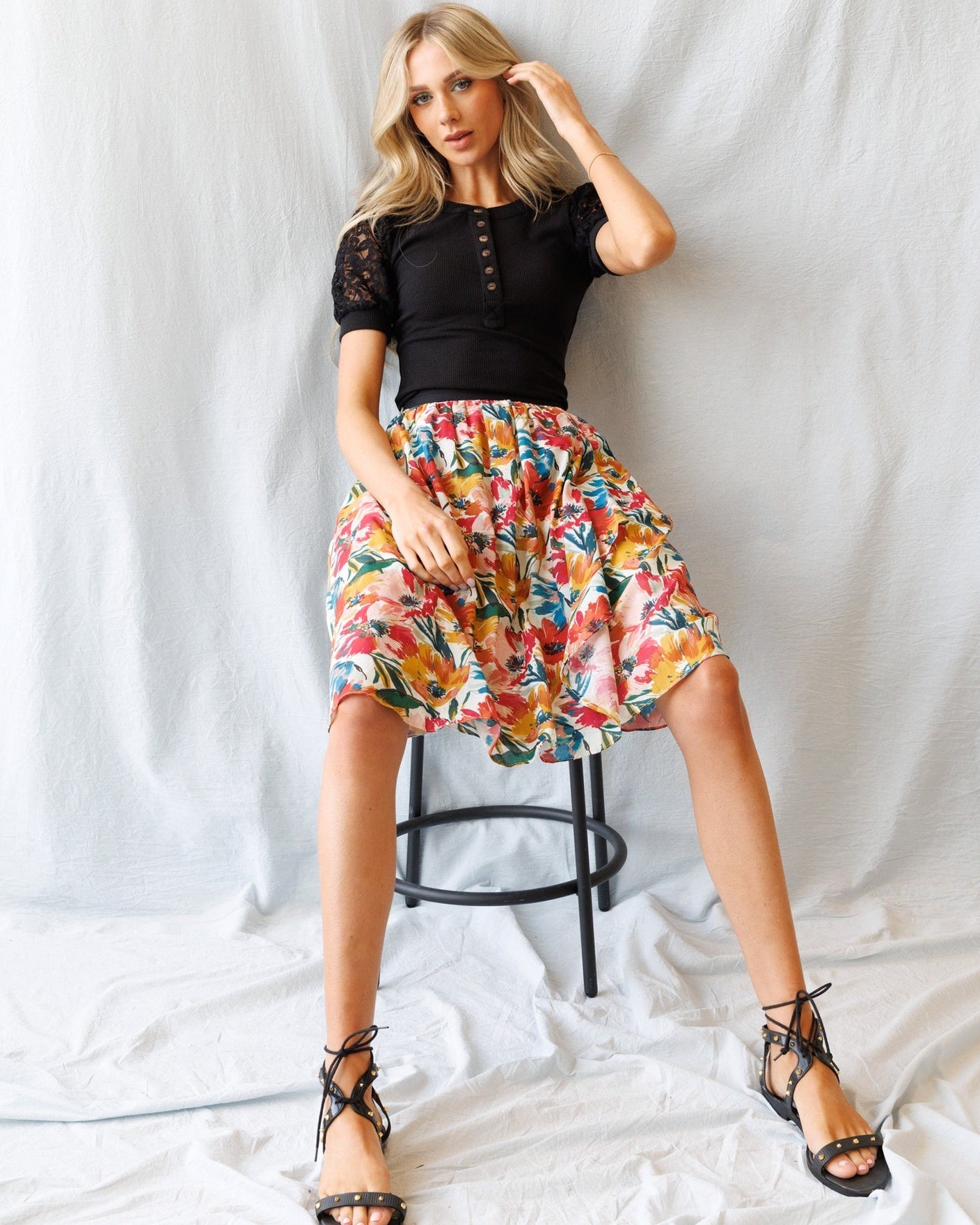 Woman sitting on a stool wearing a black top and colorful floral skirt against a white background