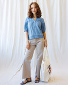 Woman wearing a blue top and light gray pants, holding a white tote bag with a design, against a white curtain background.