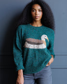 A woman with long, dark curly hair wearing a teal green fuzzy knit sweater with a large seagull motif across the chest, paired with blue jeans, standing against a dark blue-gray paneled wall.
