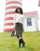 A woman stands on grass in front of a red-and-white striped lighthouse and white building, wearing a cream long-sleeve top, olive green skirt, dark green rain boots, and holding a blue-and-white patterned garment and a small white bag.