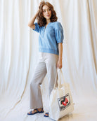 Woman wearing a blue top and light gray pants, holding a white tote bag with a design, against a white curtain background.