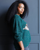A woman with long, curly black hair is shown from the side, wearing a fuzzy teal-green knit sweater with a white and gray geometric pattern on the front, posed against a dark blue paneled wall.