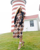 A woman stands on a grassy lawn in front of a red-and-white striped lighthouse and a white building, wearing a long patterned midi dress with a black cardigan and beige ankle boots.