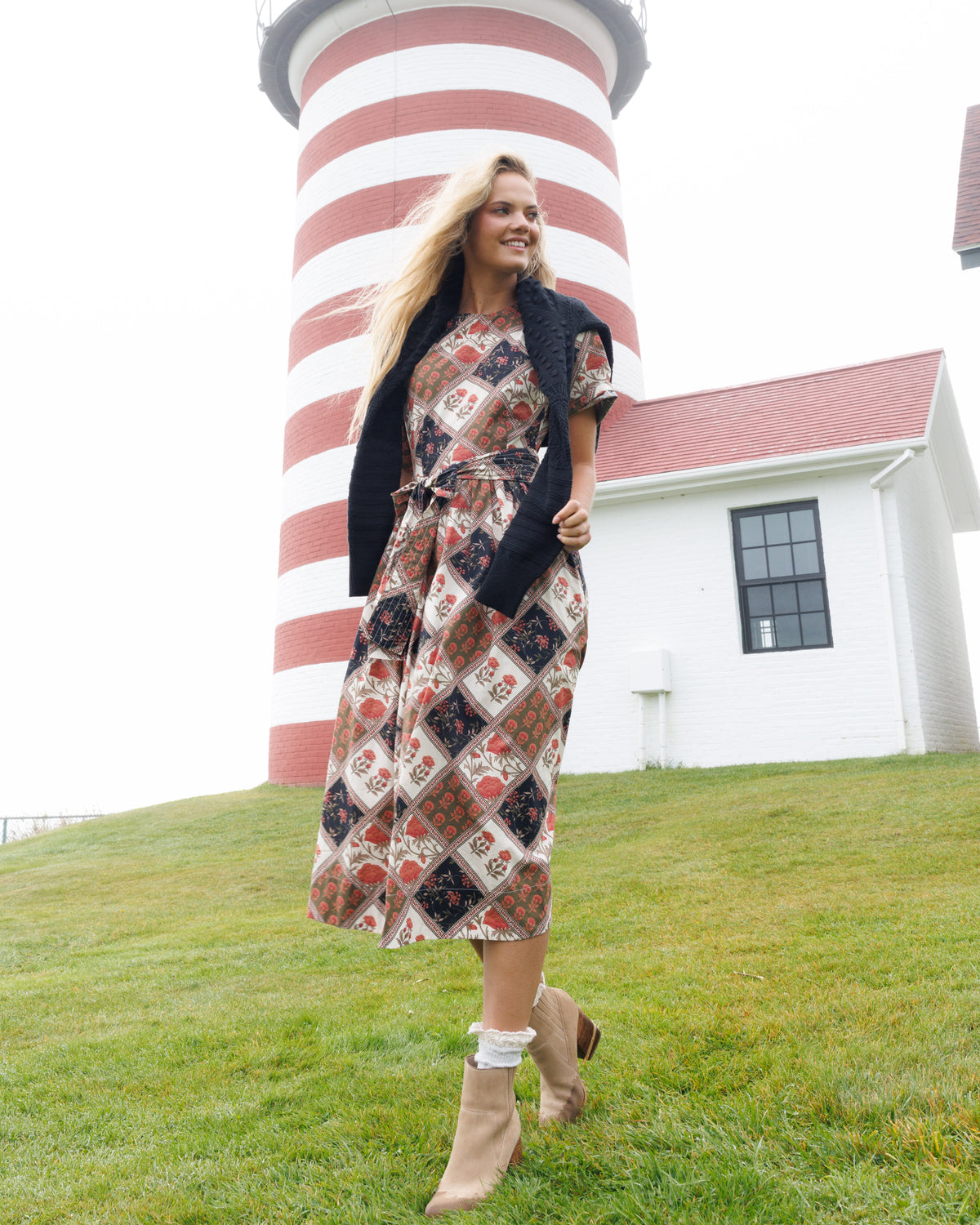 A woman stands on a grassy lawn in front of a red-and-white striped lighthouse and a white building, wearing a long patterned midi dress with a black cardigan and beige ankle boots.