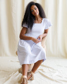 Woman wearing a purple checkered dress sitting on a white surface with a white curtain background
