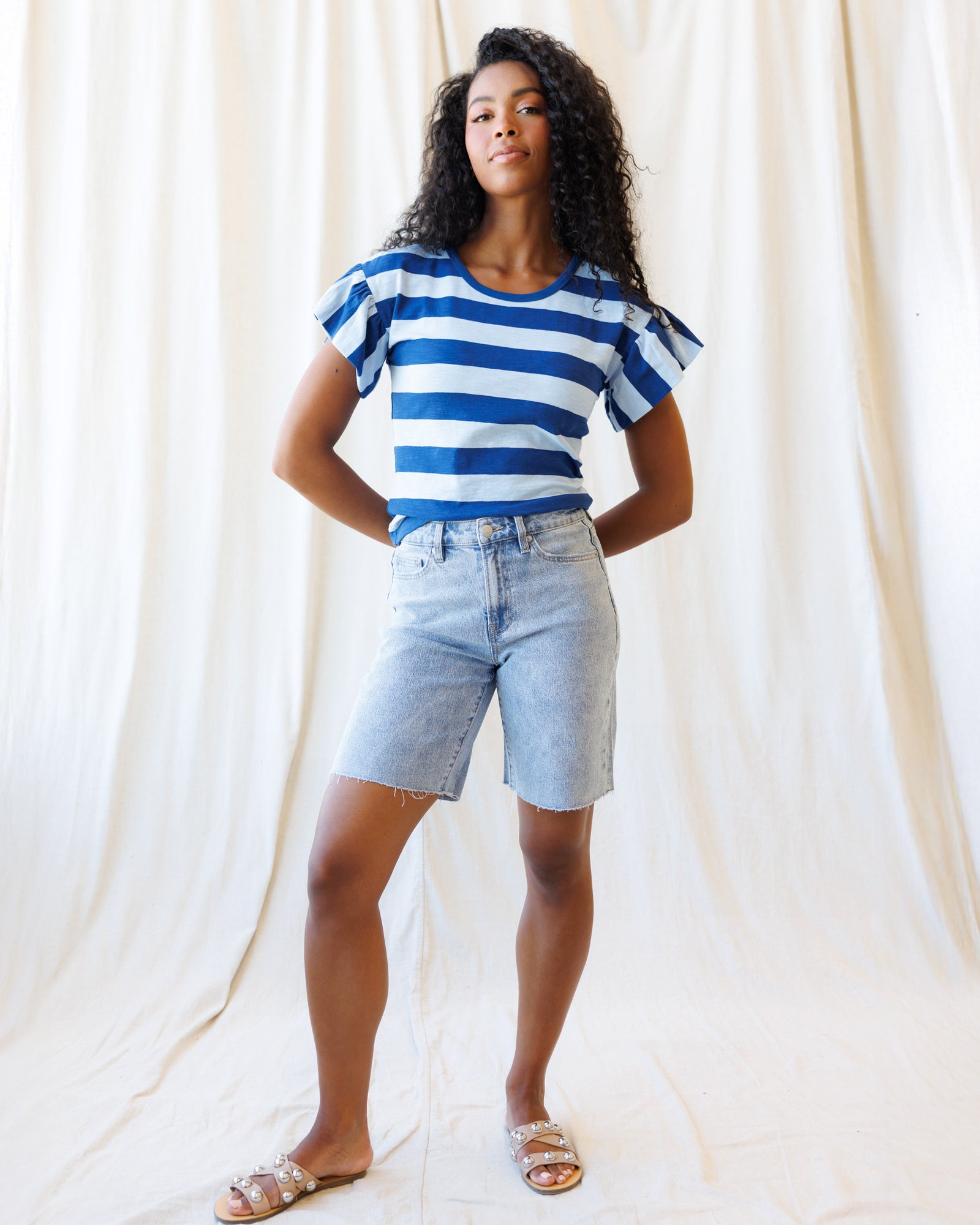 Woman wearing a blue and white striped shirt and denim shorts against a white curtain background