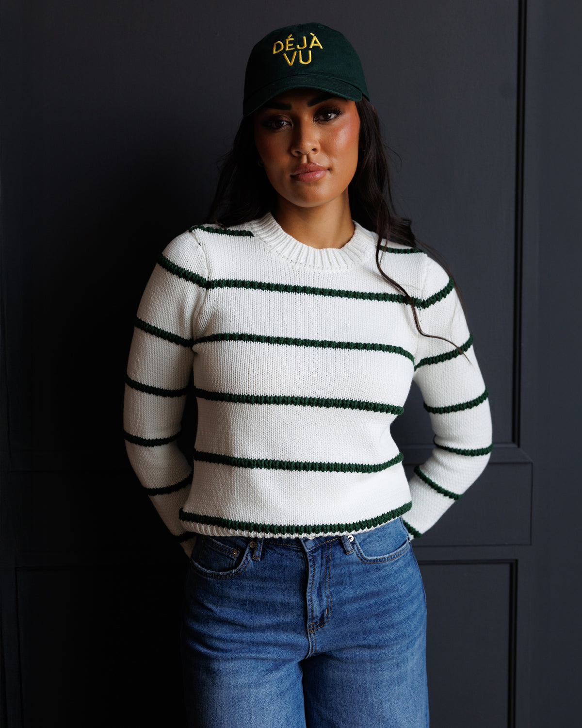 A woman wearing a white knit sweater with dark green horizontal stripes, blue jeans, and a dark green baseball cap with yellow text, standing against a dark gray paneled wall.