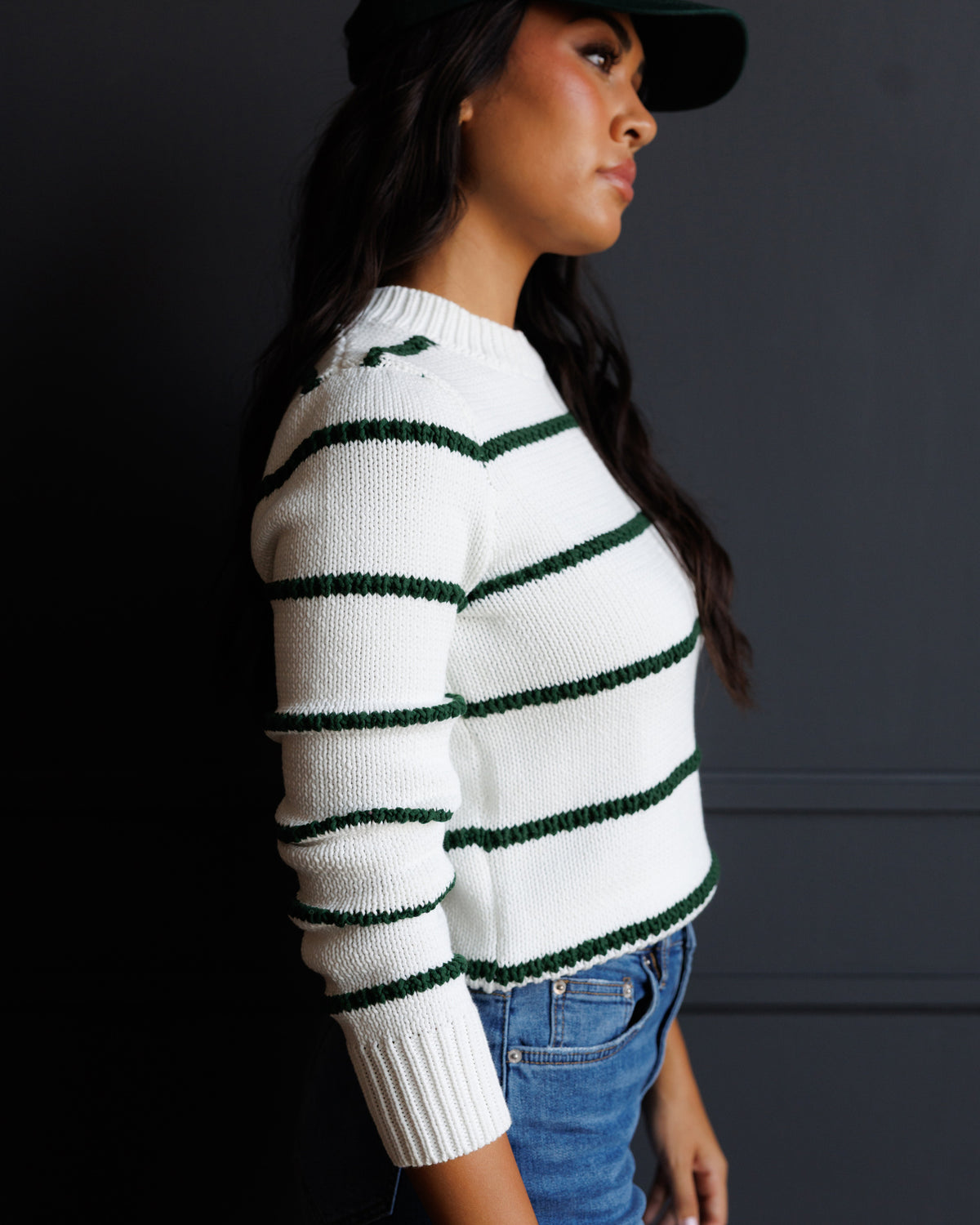 A woman in profile wearing a white knit sweater with dark green horizontal stripes, paired with blue jeans and a black cap, set against a dark gray background.