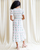 Woman wearing a white dress with green floral pattern against a white curtain background