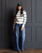 A woman stands against a dark paneled wall wearing a cream knit sweater with dark green horizontal stripes, blue wide-leg jeans, a dark green baseball cap with yellow lettering, and leopard-print sandals.