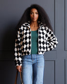 A woman with curly black hair stands against a dark gray paneled wall wearing a black and white diamond-patterned zip-up jacket over a green top and light blue jeans.
