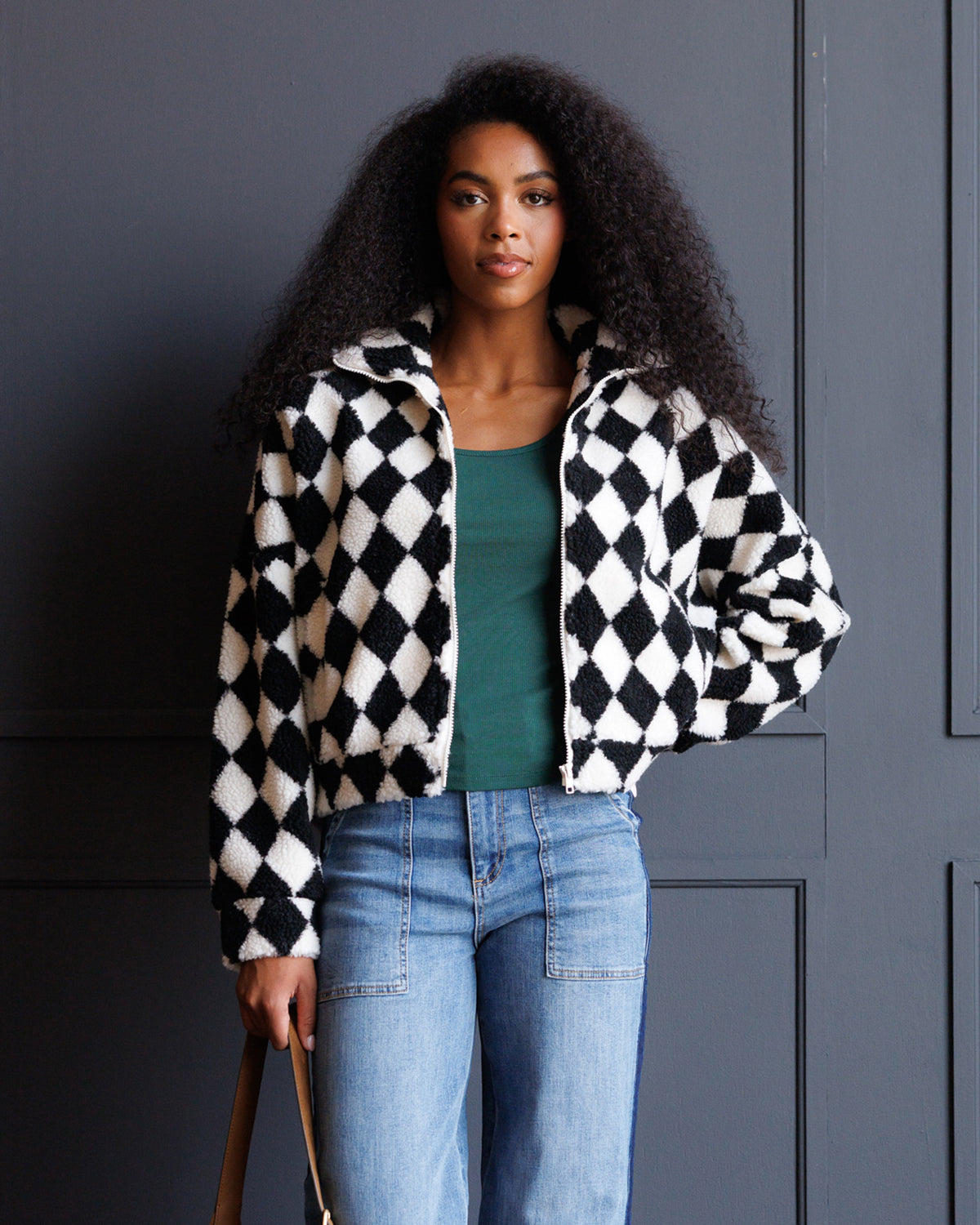A woman with curly black hair stands against a dark gray paneled wall wearing a black and white diamond-patterned zip-up jacket over a green top and light blue jeans.