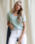Woman wearing a light green crochet top and white pants against a neutral background