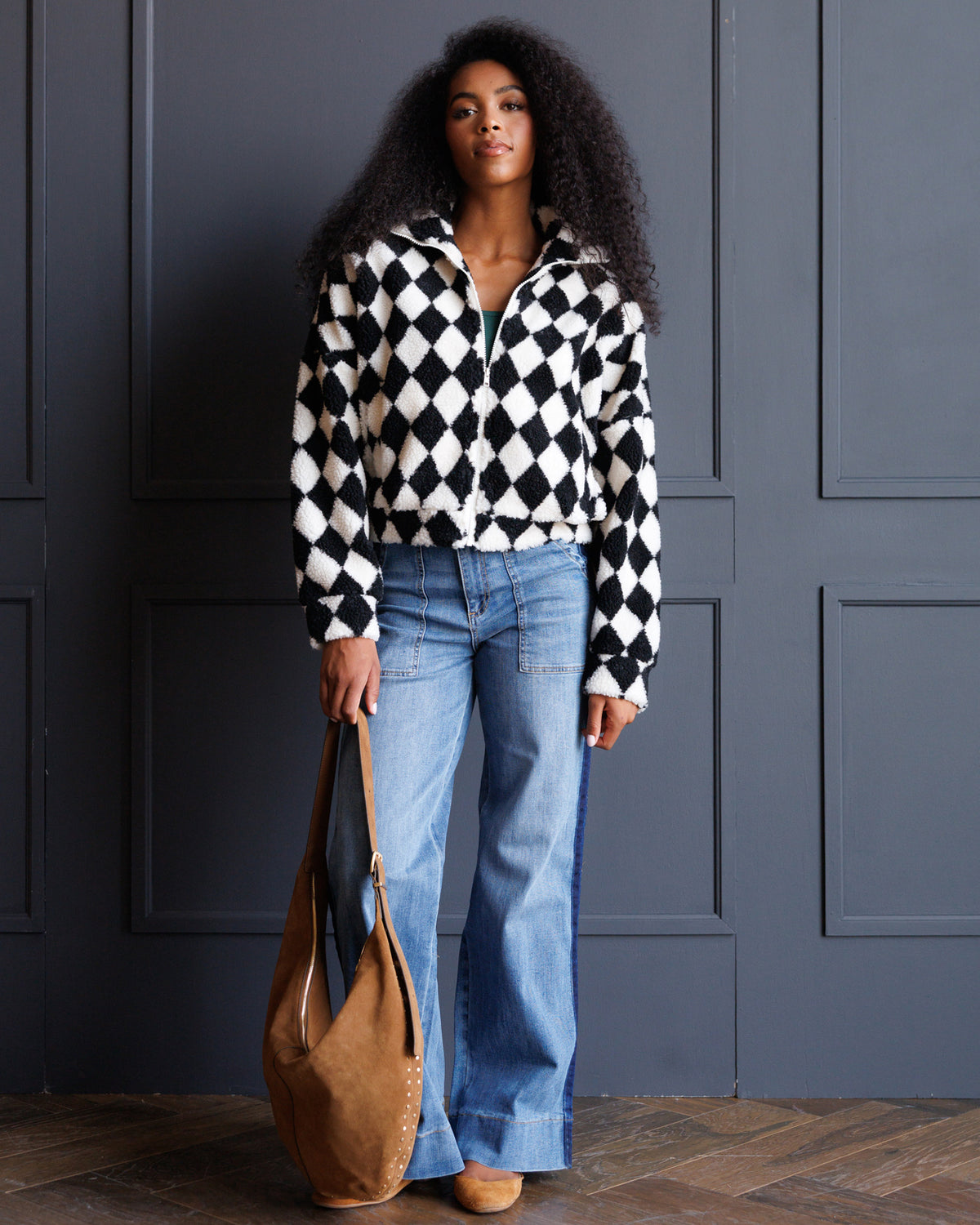 A woman stands against a dark gray paneled wall wearing a black and white harlequin diamond-patterned zip-up jacket, light blue wide-leg jeans, and carrying a large tan shoulder bag.