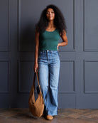 A woman with long curly black hair stands against a dark blue paneled wall wearing a dark green tank top, high-waisted light blue wide-leg jeans, and tan shoes, while holding a large tan suede shoulder bag.