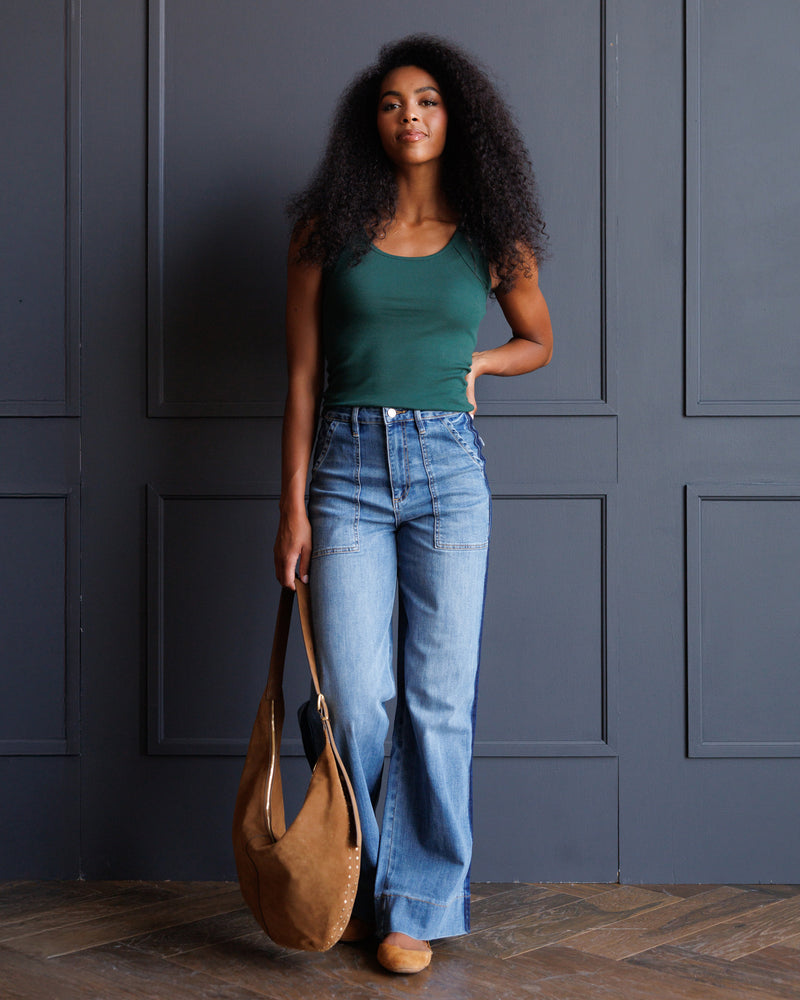A woman with long curly black hair stands against a dark blue paneled wall wearing a dark green tank top, high-waisted light blue wide-leg jeans, and tan shoes, while holding a large tan suede shoulder bag.