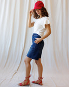 Woman wearing a white t-shirt, blue denim skirt, red cap, and sandals against a plain background