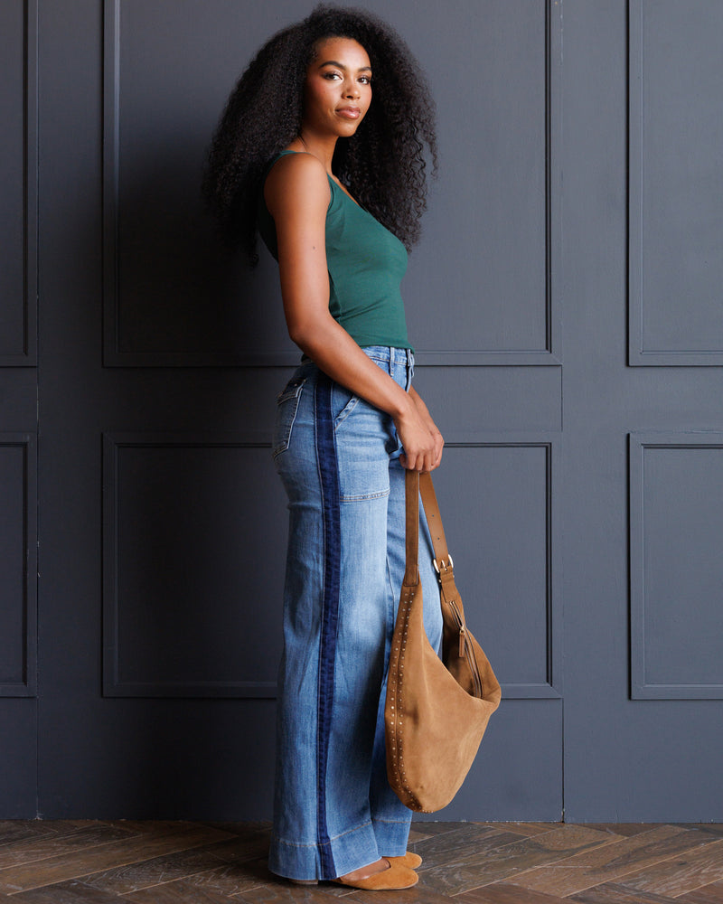 A woman stands in profile against a dark gray paneled wall, wearing a green tank top, wide-leg blue jeans, and tan shoes, while holding a tan suede shoulder bag.