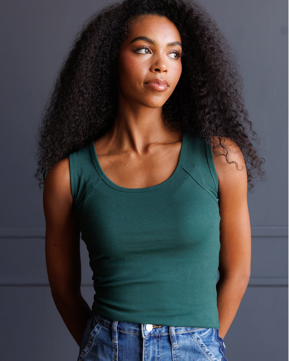 A woman with long, curly black hair wearing a fitted dark green scoop-neck tank top and blue jeans, standing against a dark gray paneled wall.