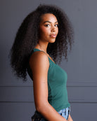 A woman with voluminous dark curly hair is shown in profile wearing a fitted teal-green tank top and light-wash denim jeans against a dark gray paneled wall.