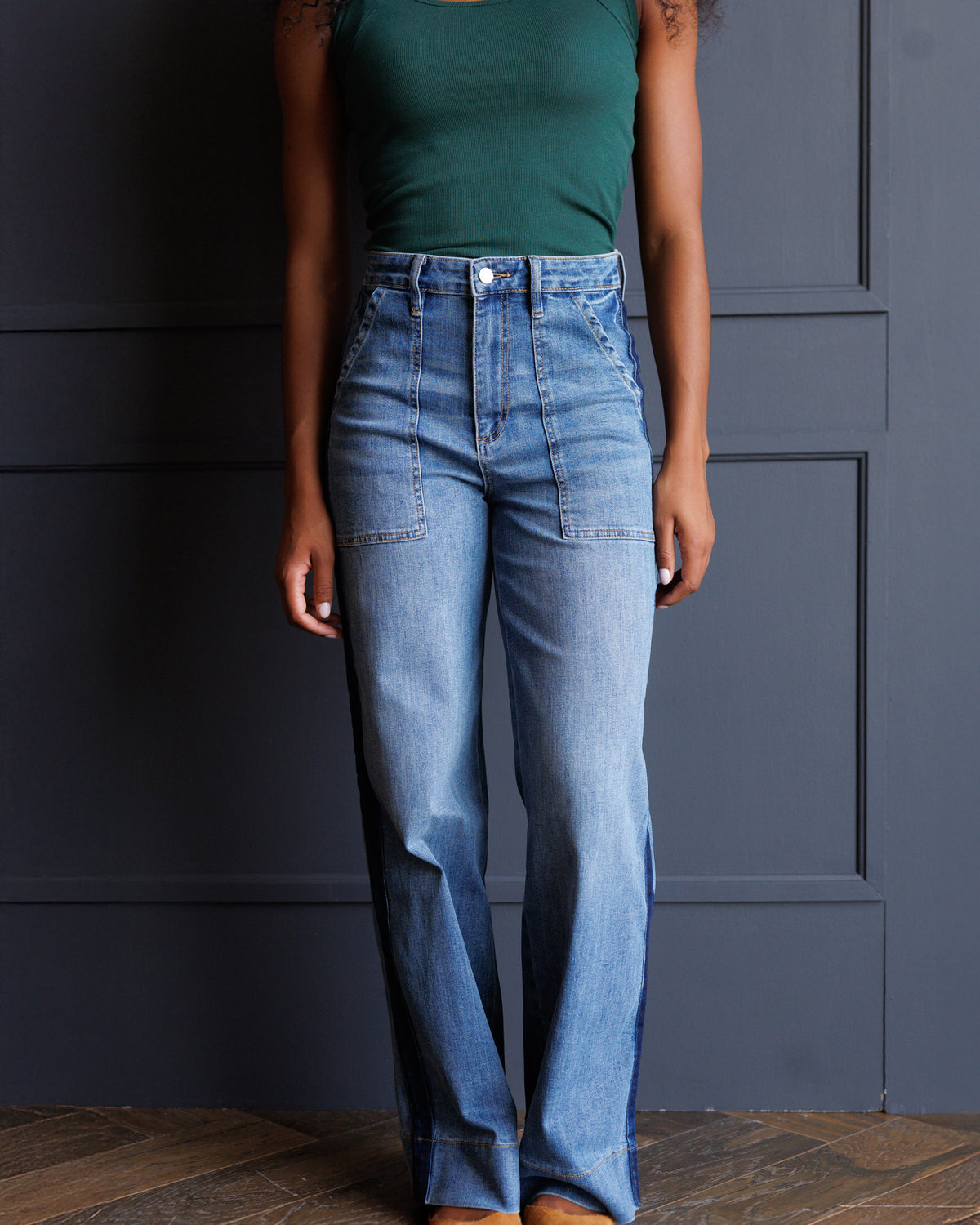 A model wearing high-waisted, wide-leg light blue denim jeans with front patch pockets, paired with a dark green tank top, standing against a dark gray paneled wall.