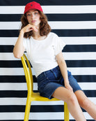 Woman sitting on a yellow chair wearing a white t-shirt and denim shorts against a black and white striped background.