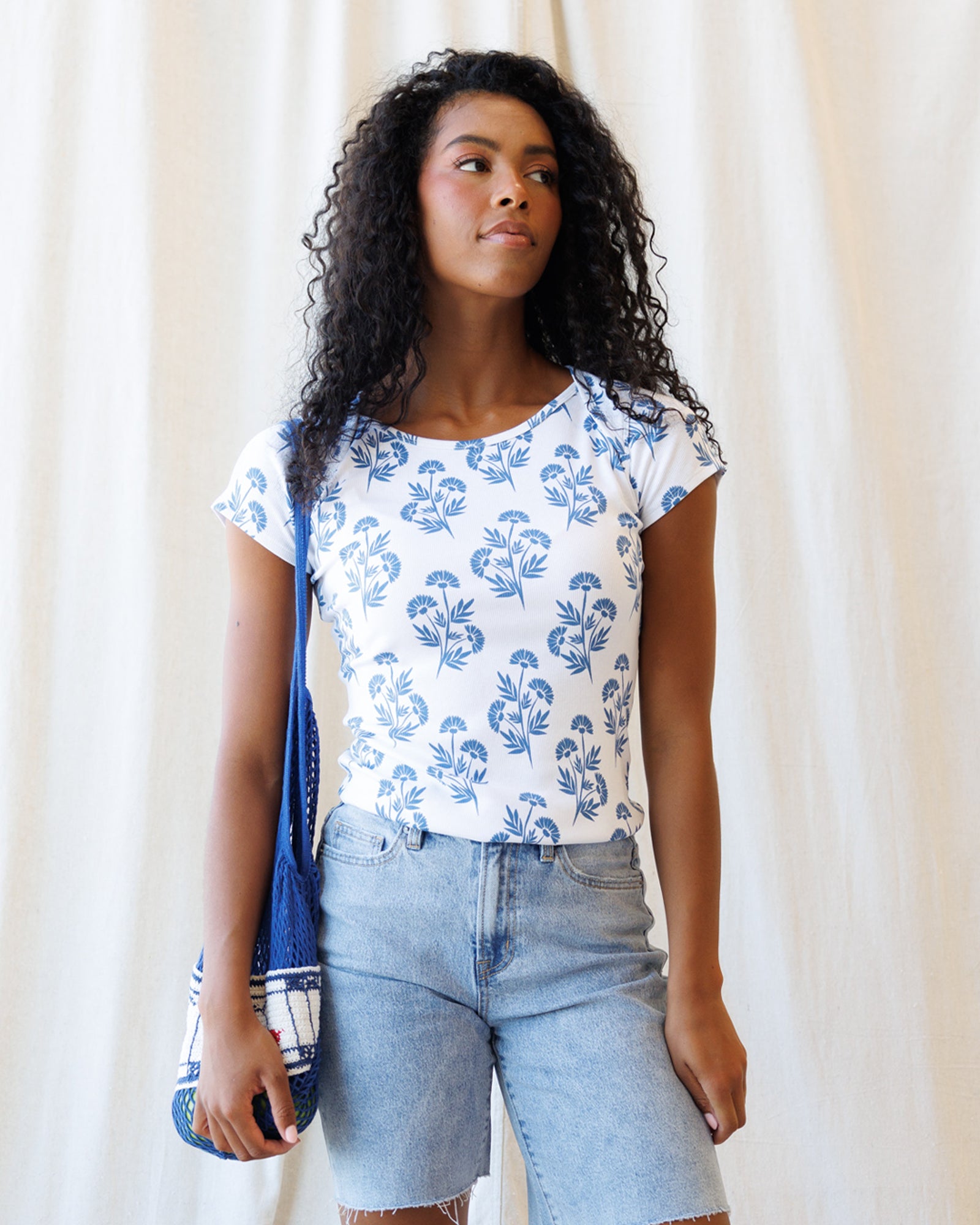 Woman wearing a white t-shirt with blue floral pattern and light blue shorts, holding a blue bag against a white curtain background.