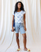 Woman wearing a floral top and denim shorts holding a blue bag against a white curtain background