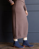 A person wearing a ribbed knit midi skirt in a muted taupe-brown color, paired with navy blue suede ankle boots and cream socks, standing against a dark blue paneled wall.