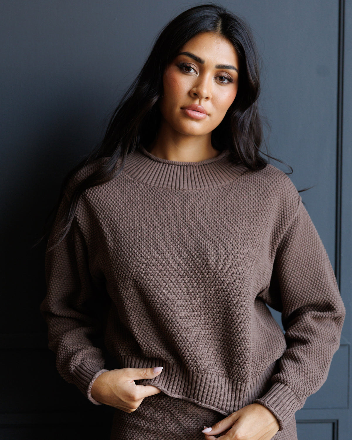 A woman wearing a textured brown knit crewneck sweater, posing against a dark gray paneled wall.