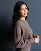 A woman with long dark hair wearing a textured brown knit sweater, posing against a dark gray wall.