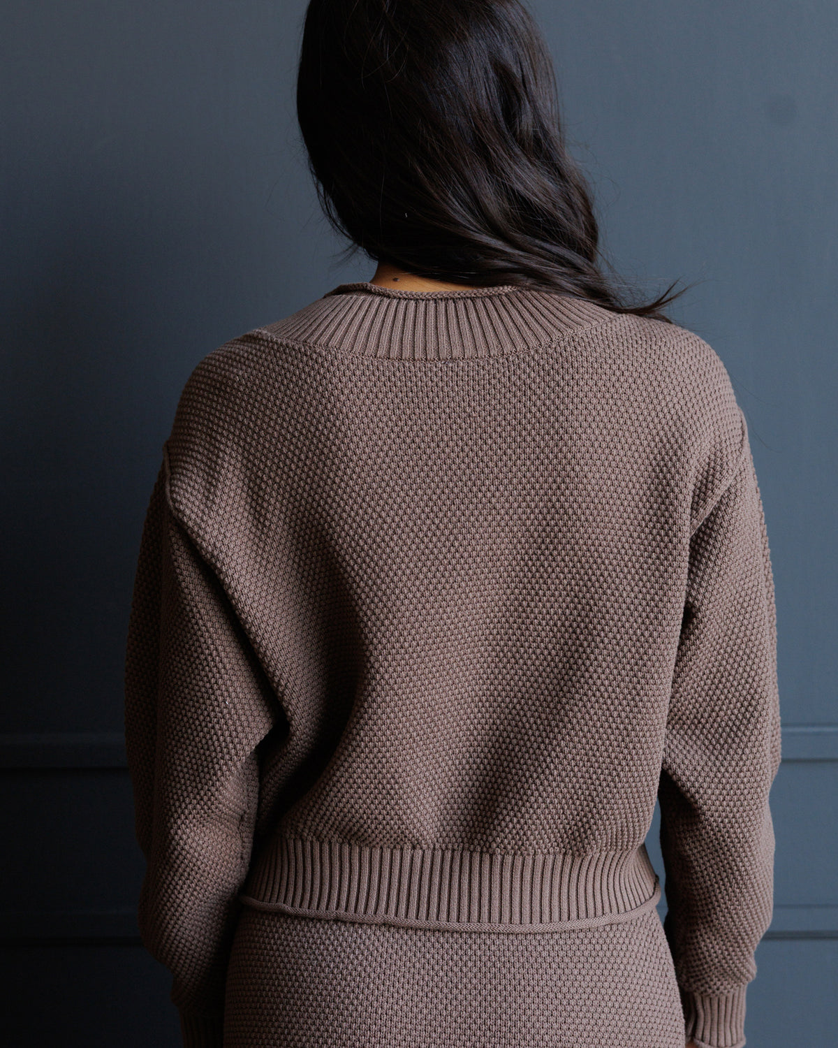 A person shown from the back wearing a textured knit sweater in a muted taupe-brown color, set against a dark gray wall.