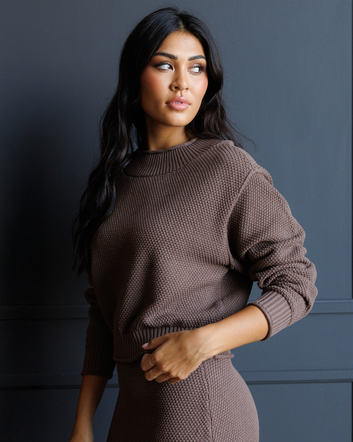 A woman wearing a textured brown knit mock-neck sweater and matching skirt, posing against a dark gray paneled wall.