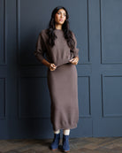 A woman models a brown knit sweater and matching midi skirt set, standing against a dark blue paneled wall.