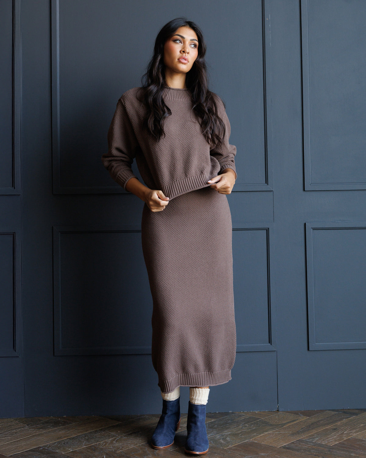 A woman models a brown knit sweater and matching midi skirt set, standing against a dark blue paneled wall.