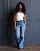 A woman with curly black hair stands against a dark blue paneled wall wearing a white sleeveless top and light blue wide-leg jeans with a black belt.